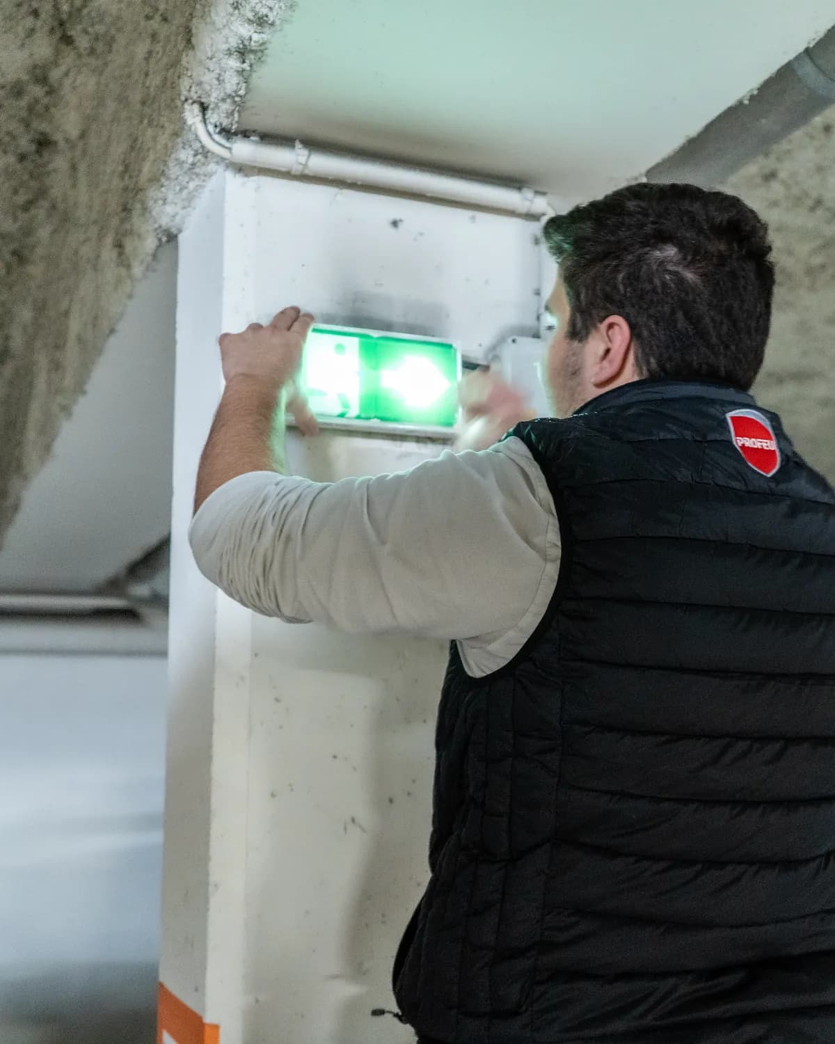 Technicien PROFEU vérifiant un bloc autonome d'éclairage de sécurité (BAES) dans un parking souterrain